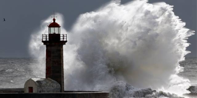 Roter Leuchtturm auf einer Mole, von einer gewaltigen, brechenden Welle umspült.